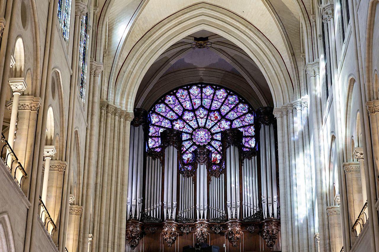 Uma vista interna da catedral mostra janelas e vigas ornamentadas, com um vitral roxo na frente, acima dos muitos tubos de prata do órgão restaurado.