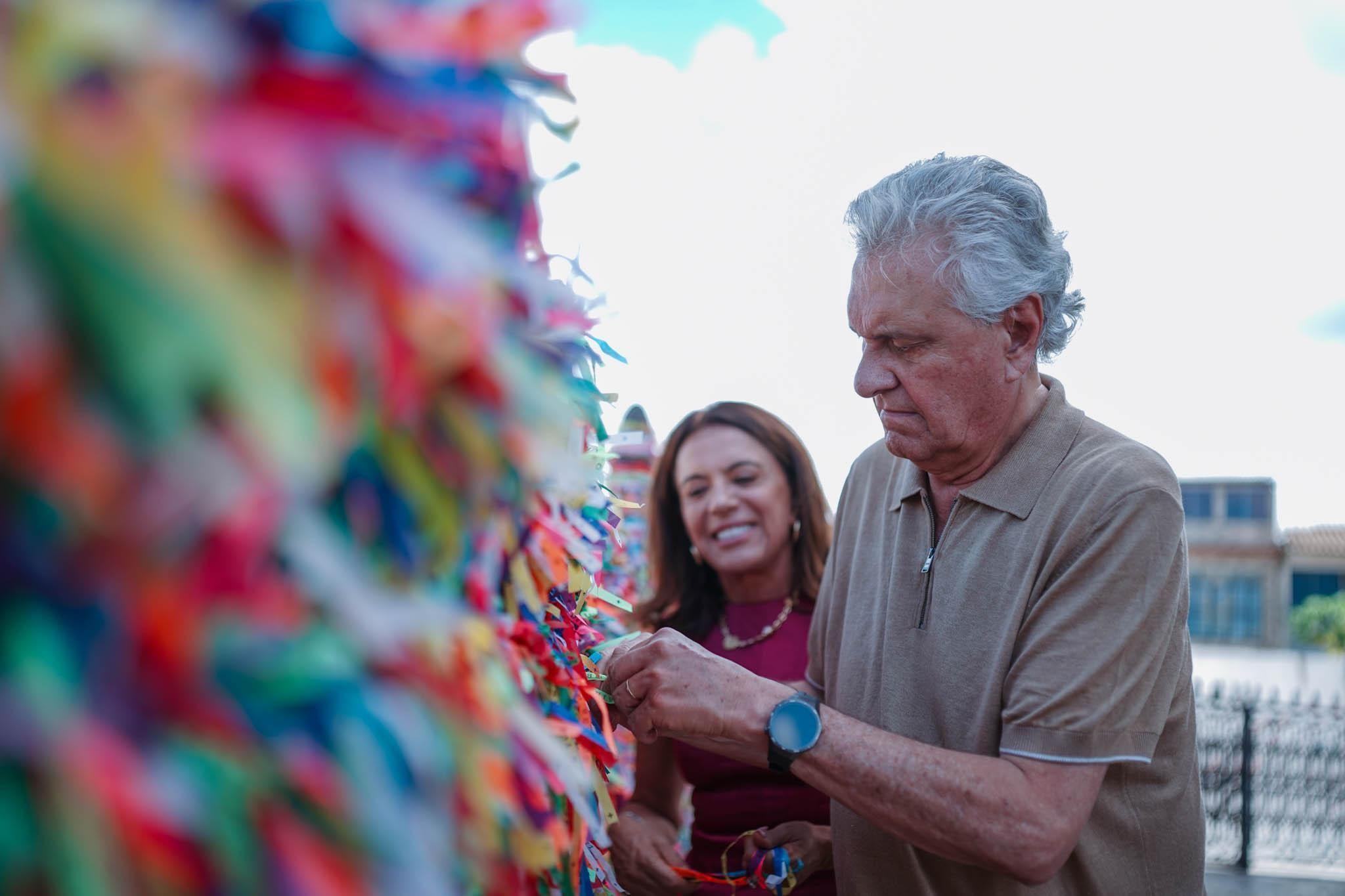 Caiado mexendo em fitinhas do Senhor do Bonfim, com esposa atrás, sorrindo