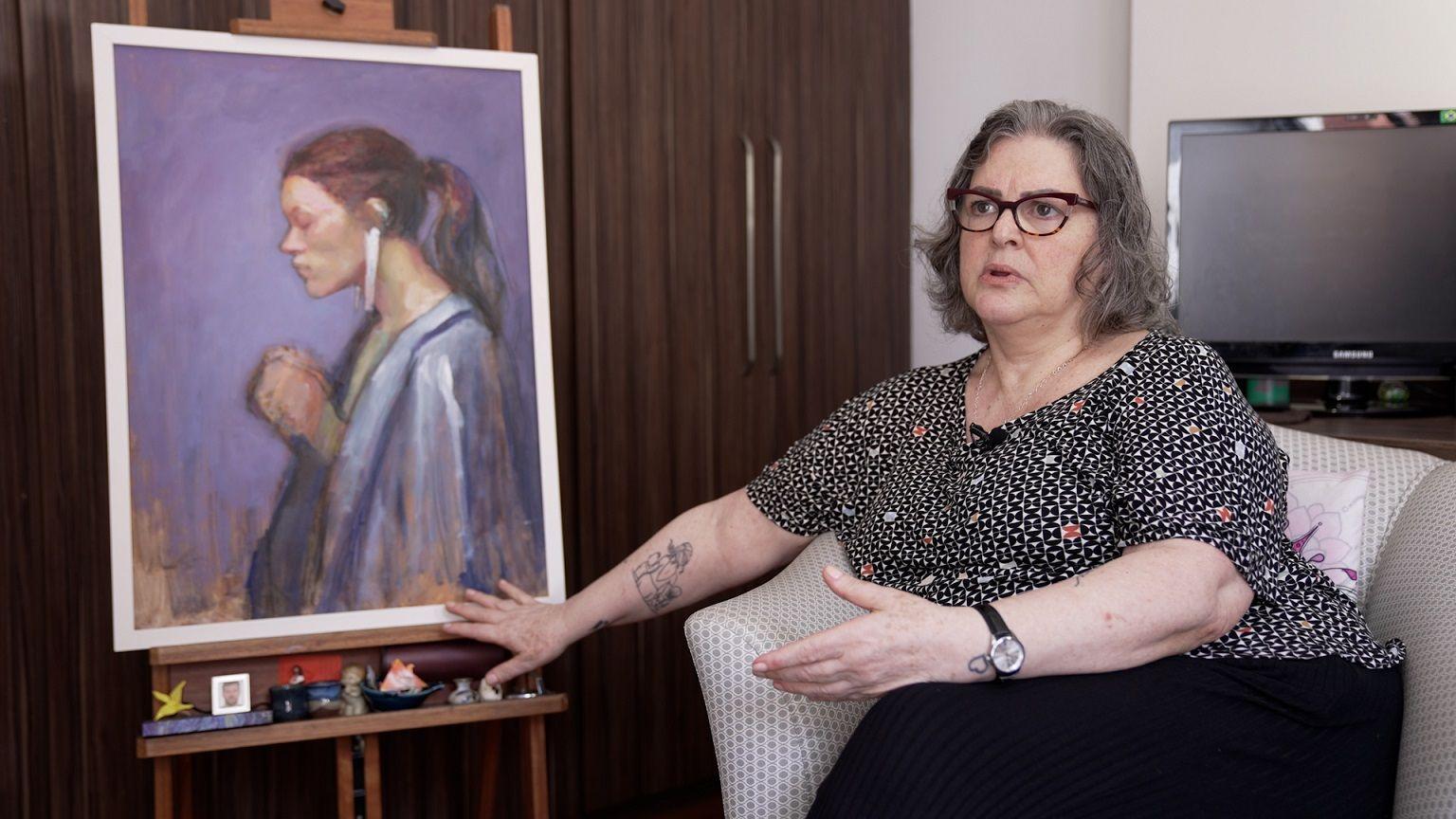 Maria Cecilia sentada e conversando ao lado de pintura de uma mulher