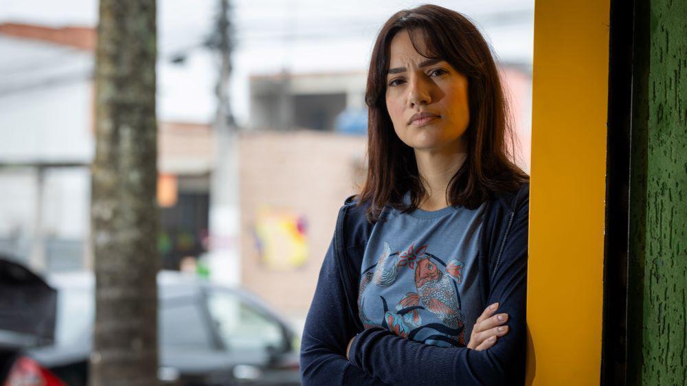 Foto de Patrícia, uma mulher jovem, de pele clara, cabelo liso e escuro, vestindo uma blusa azul de manga cumprida, de braços cruzados e semblante sério.