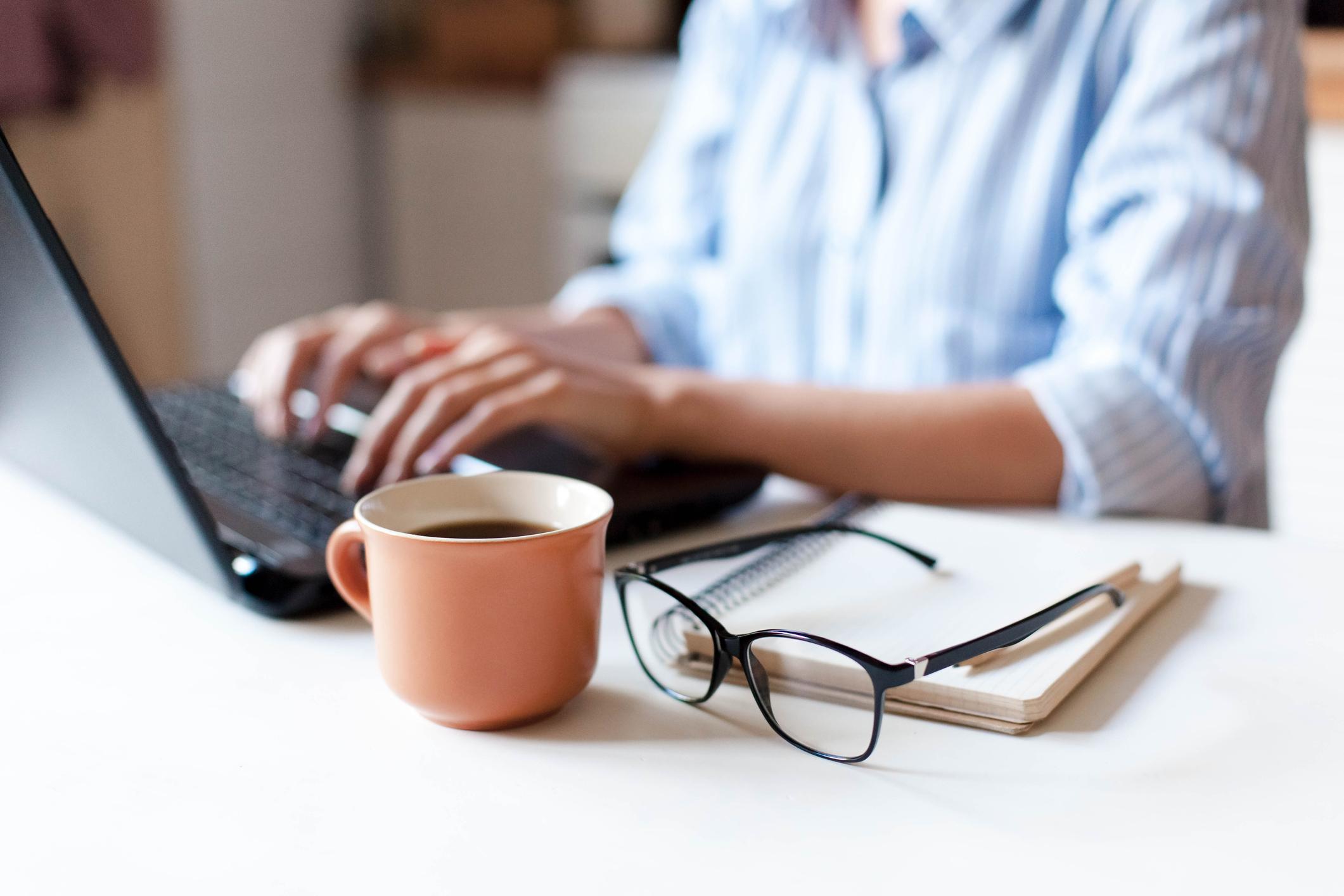 Pessoa digitando no laptop com xícara de café e óculos sobre a mesa