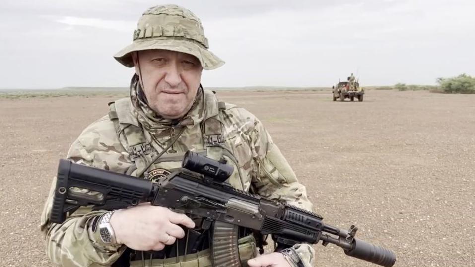 Yevgeny Prigozhin em traje militar camuflado, segurando uma arma, em paisagem desértica com caminhonete ao fundo, em vídeo compartilhado em agosto de 2023.