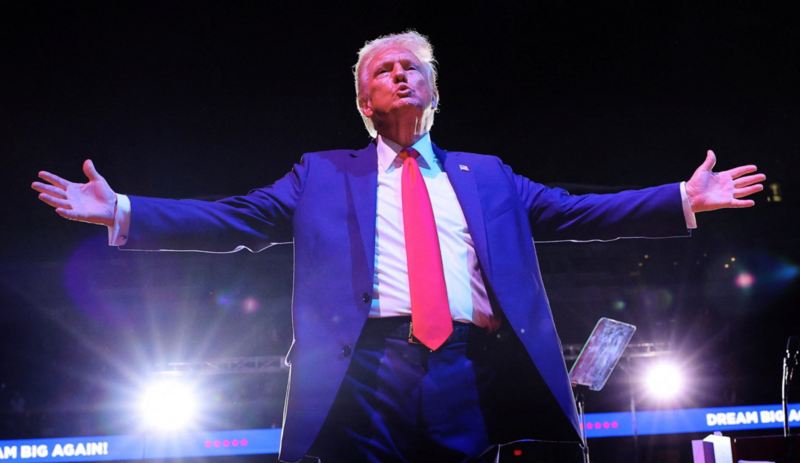 Trump abrindo os braços em uma arena iluminada durante um grande evento