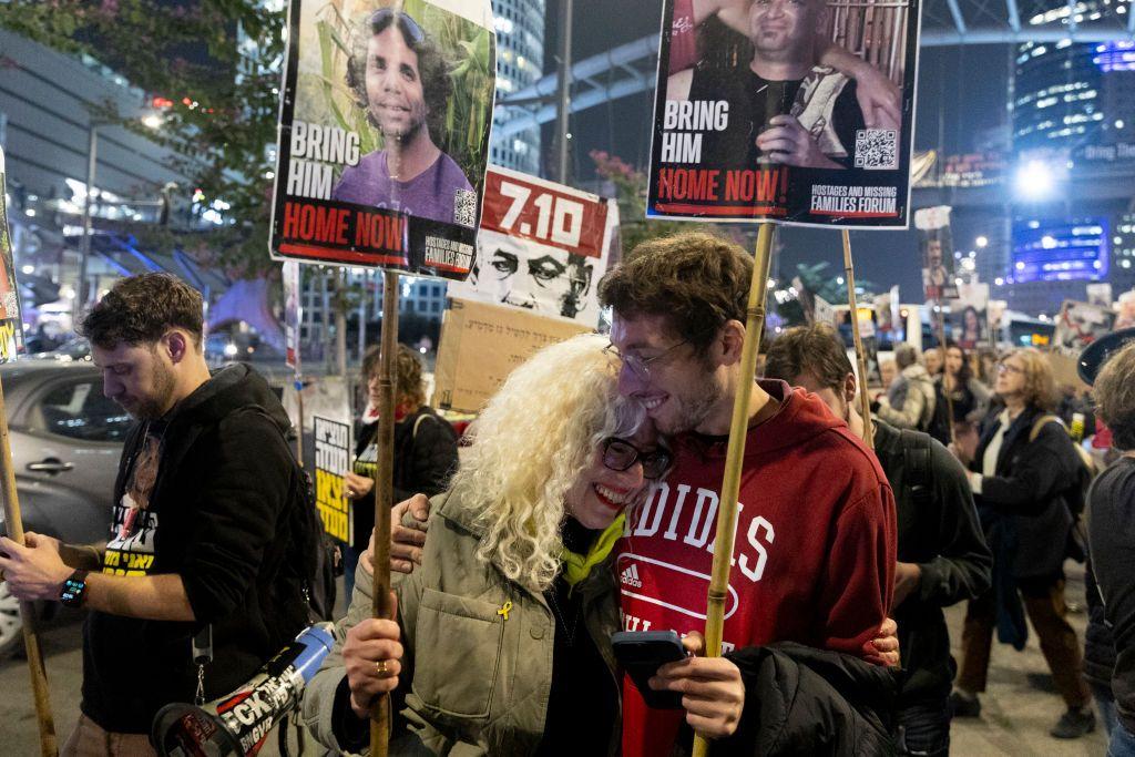 Duas pessoas se abraçando e sorrindo em rua durante a noite, com mais manifestantes atrás