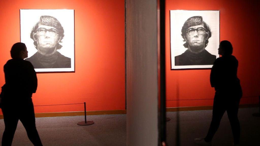 A woman looking at the piece 'Keith Mezzotint’ by Chuck Close, and a reflection of her and the piece are seen in the mirror 