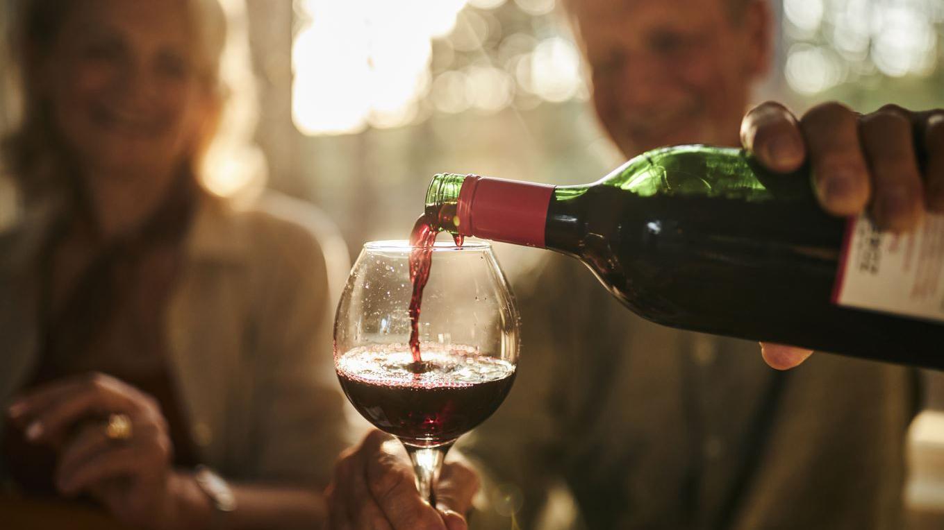 Homem sorrindo ao colocar vinho em taça; e ele está ao lado de uma mulher; eles sorriem 