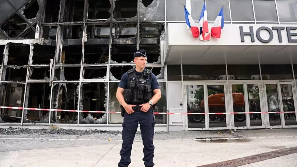 Policial com a mão na cintura na frente da fachada incendiada e depredada da prefeitura de Gonesse