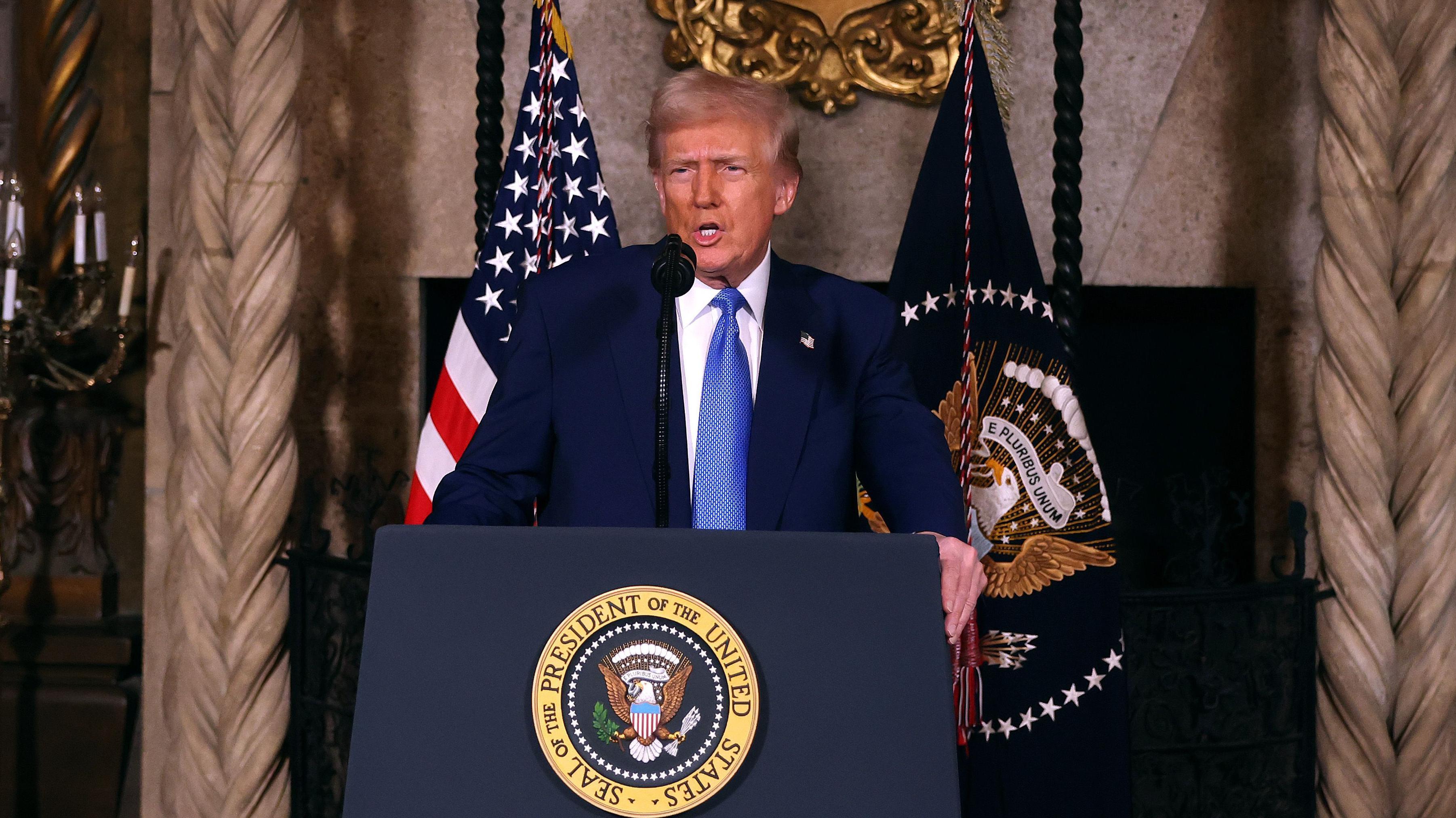 El presidente Donald Trump habla con los periodistas desde un atril con el sello del presidente de Estados Unidos. Detrás de él hay una bandera estadounidense y otra bandera con el sello del presidente. Lleva un traje oscuro y una corbata azul.