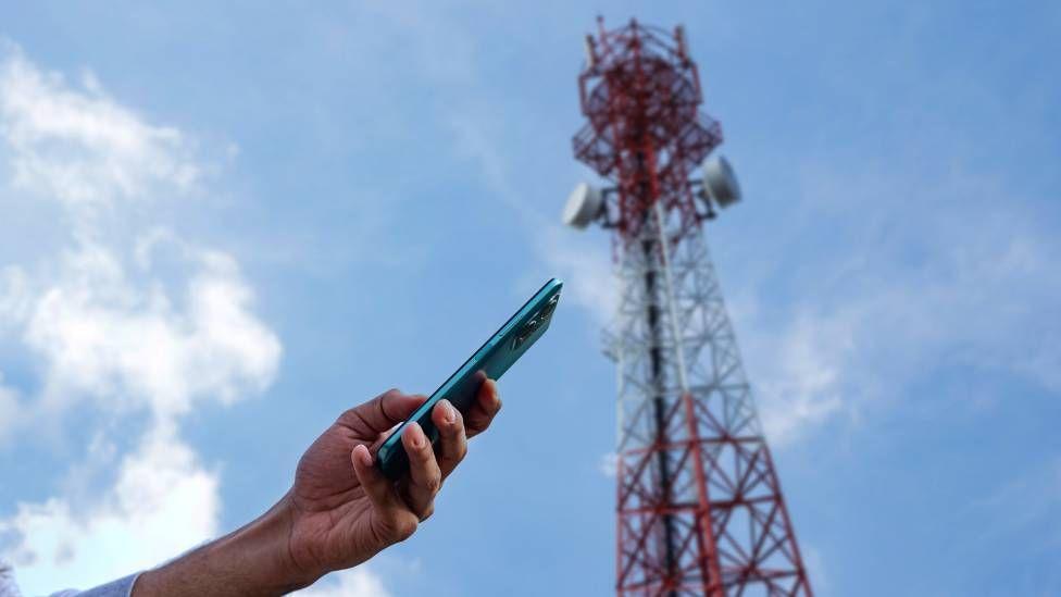 Una persona con el celular en la mano frente a una antena de telefonía móvil