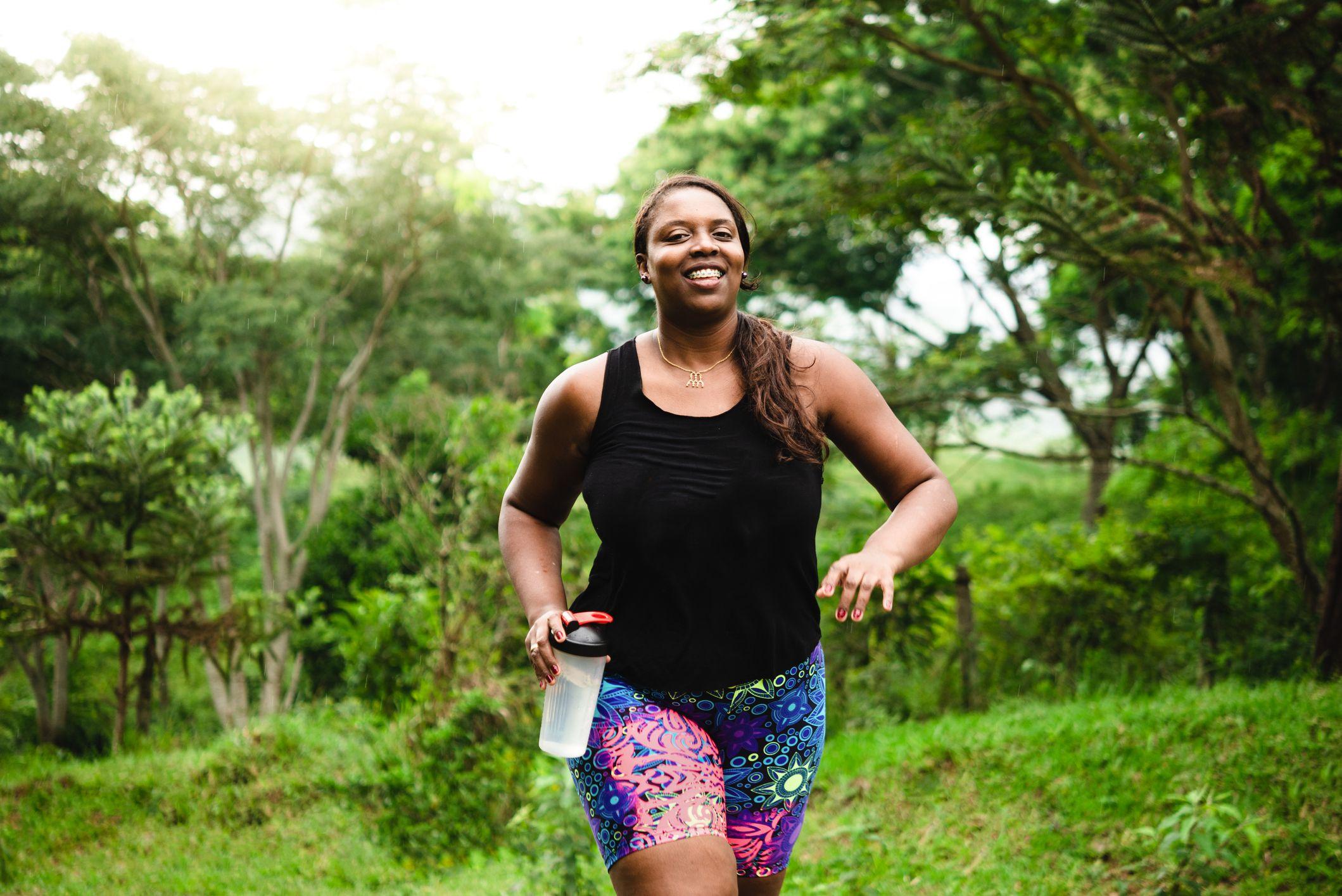 Mulher negra caminhando, sorrindo, com uma garrafa de água na mão. Ao fundo, árvores e gramado.