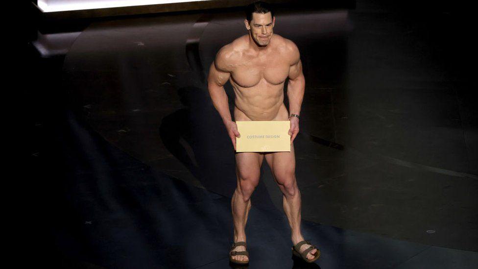John Cena sem roupas, de sandália, segura uma placa em frente à altura do quadril escrita 'figurino'
