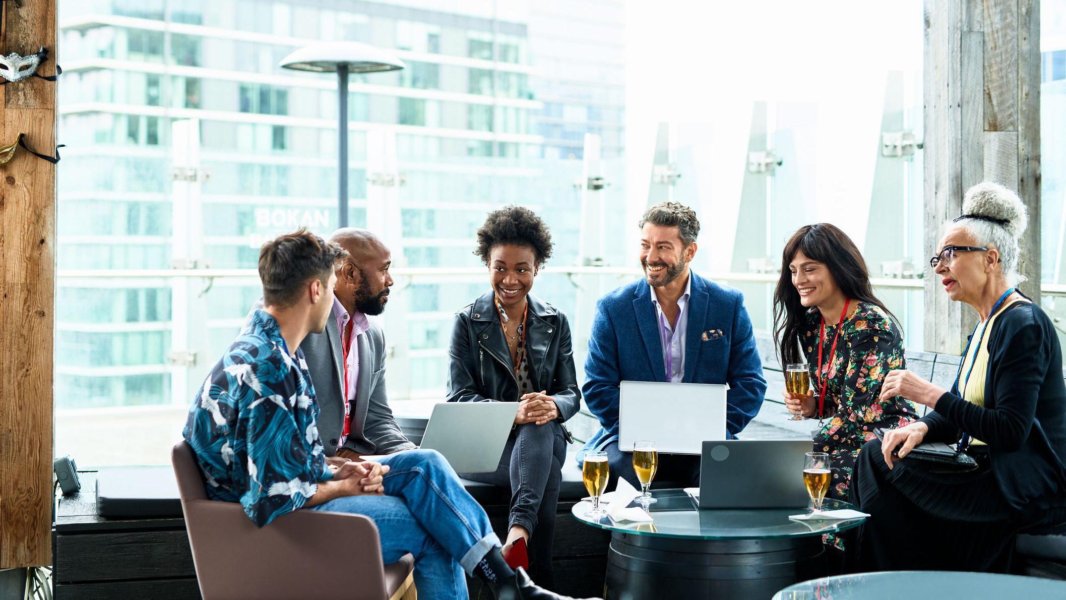 Grupo de pessoas conversando com roupas de trabalho, laptops no colo e copos de cerveja na mesa