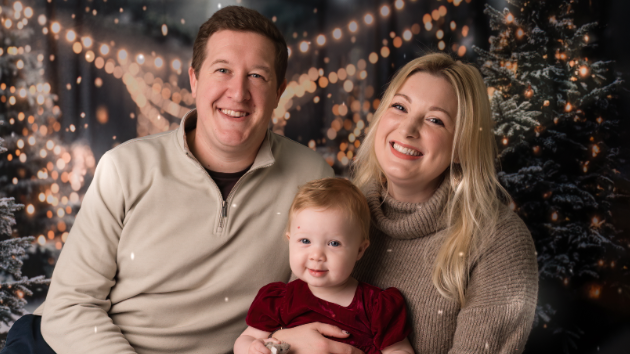 Sam e Laura sorridentes segurando a filha, Aurora, com um ano, em uma fotografia de família com árvores de Natal iluminadas ao fundo