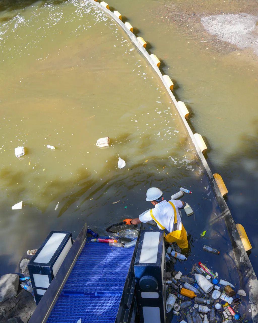 Un operario carga una cinta trasnportadora con basura plástica junto a un río.