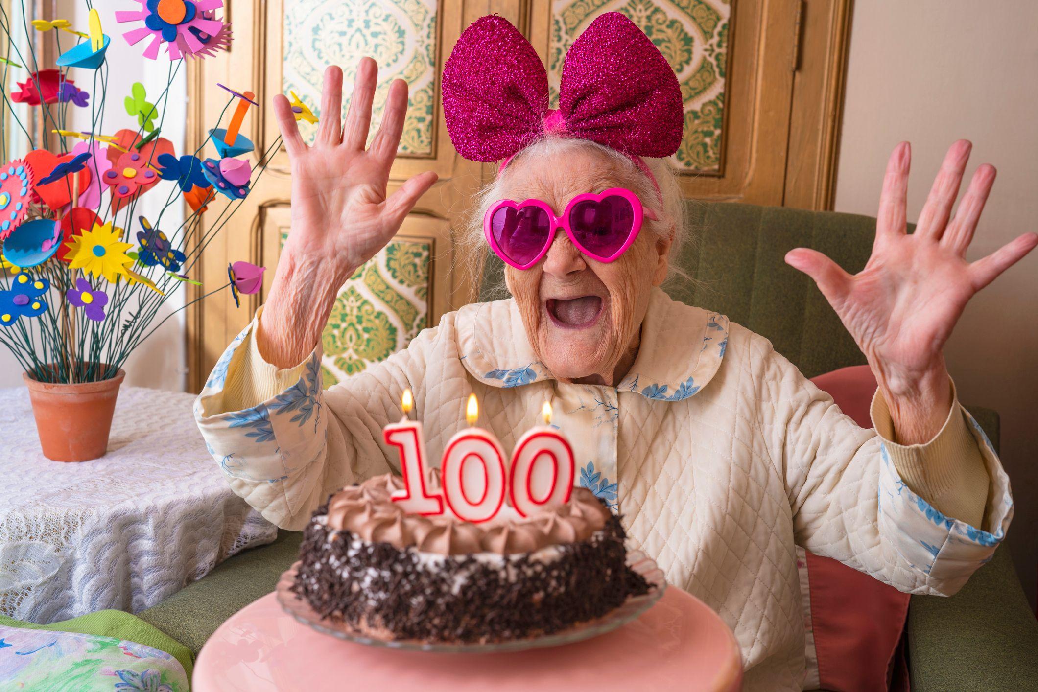 Idosa com óculos de coração e laço rosa em frnete a um bolo com velinha de 100 anos