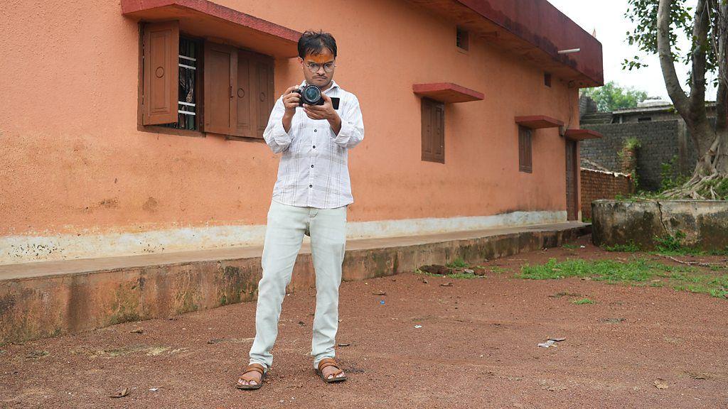 Jai Varma com roupas de cores claras e cal&ccedil;ando sand&aacute;lias, segurando uma c&acirc;mera com as m&atilde;os