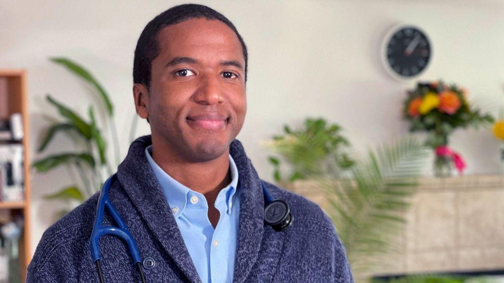 Médico com estetoscópio posa ao lado de relógio de parede em residência