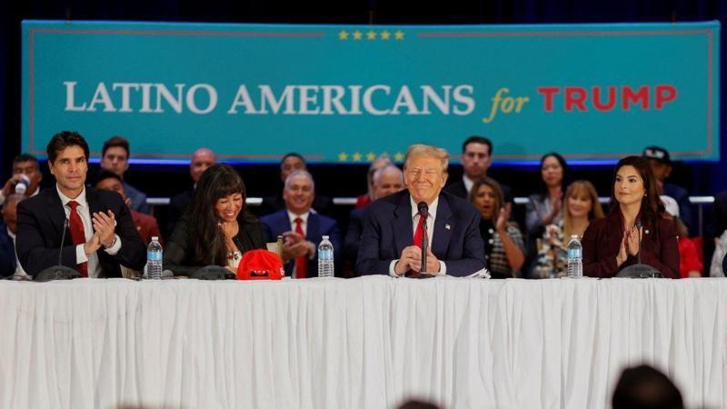 Trump no meio da mesa, sentado ao lado de diversas pessoas sorrindo; Ao fundo, uma tela diz 'Latino-Americanos por Trump'