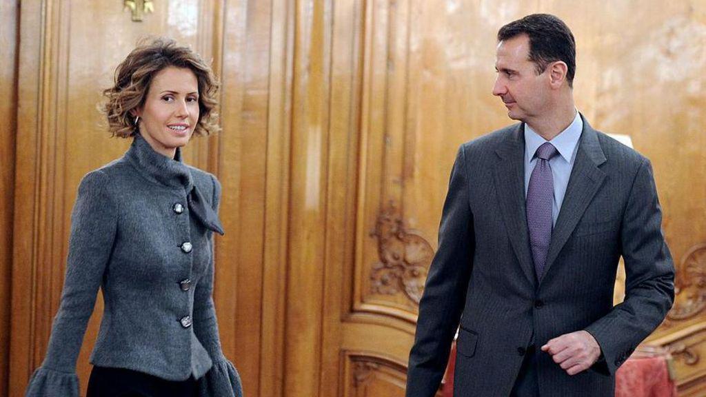 Asma al-Assad, de pelo corto y rizado, lleva una chaqueta gris y mira hacia un lado mientras su marido Bashar, con traje gris oscuro y corbata, la mira. Están de pie en una habitación revestida de madera con asientos con cojines rojos al fondo. Esta es una foto de archivo de 2010. 