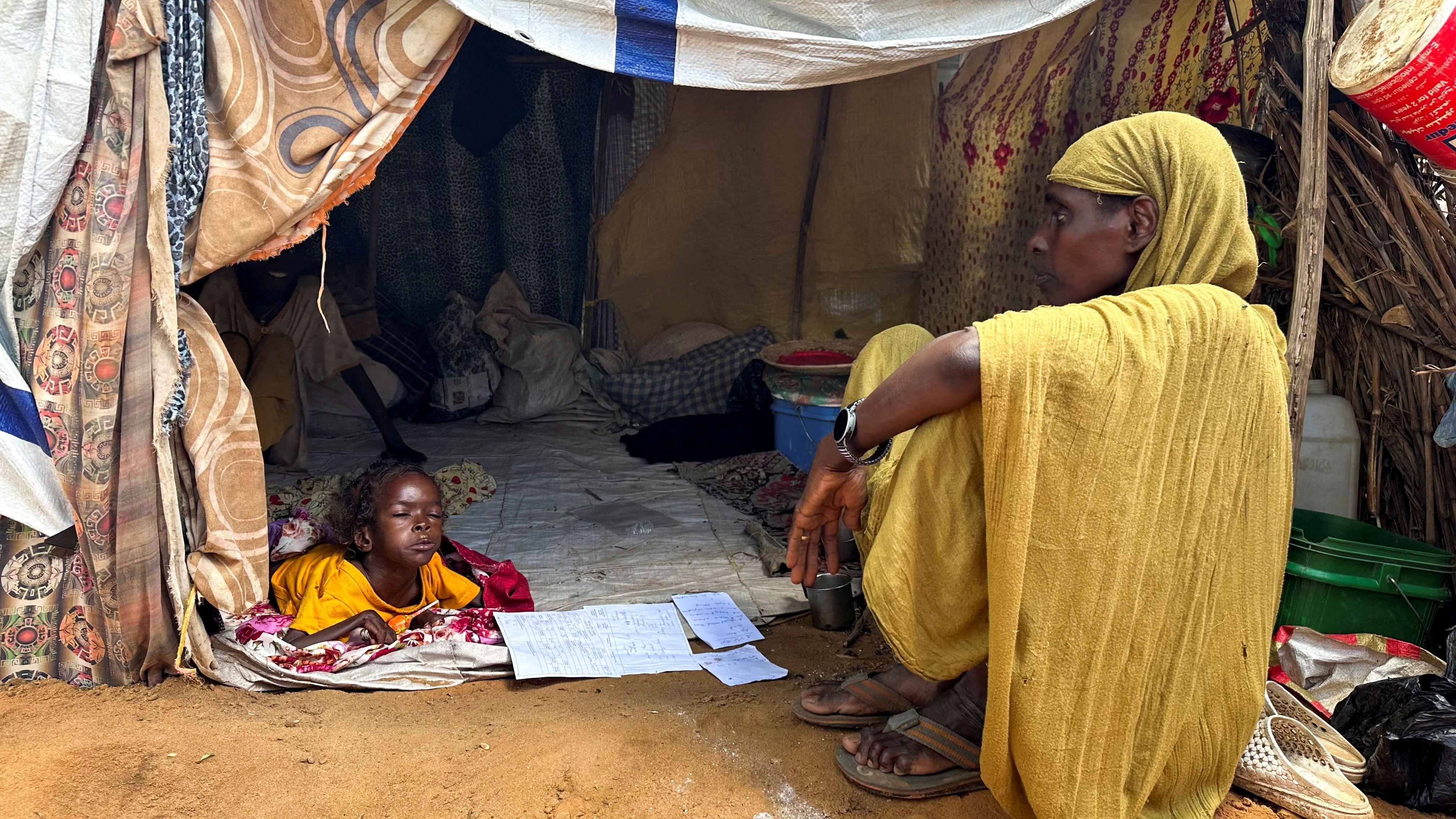 صورة لنازحة سودانية في شمال دارفور، تجلس أمام خيمة، ويبدو بداخلها طفل مستلقٍ على الأرض.