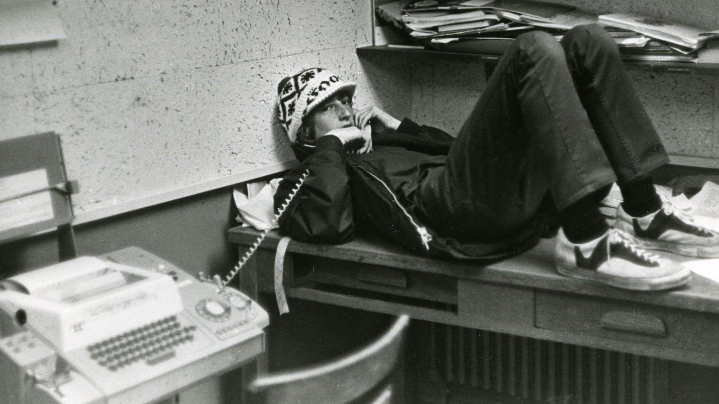 Bill Gates adolescente com chapéu, deitado sobre a mesa, encostado na parede enquanto segura um telefone fixo, em uma fotografia em preto e branco