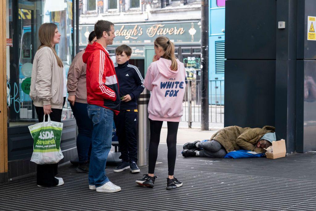 Pessoa sem-teto dorme no chão, próximo a pilastra, enquanto outras pessoas caminham com uma sacola de compras de mercado 