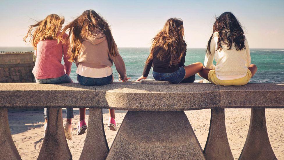 Quatro meninas sentadas de costas em mureta na praia de frente para o mar na Espanha