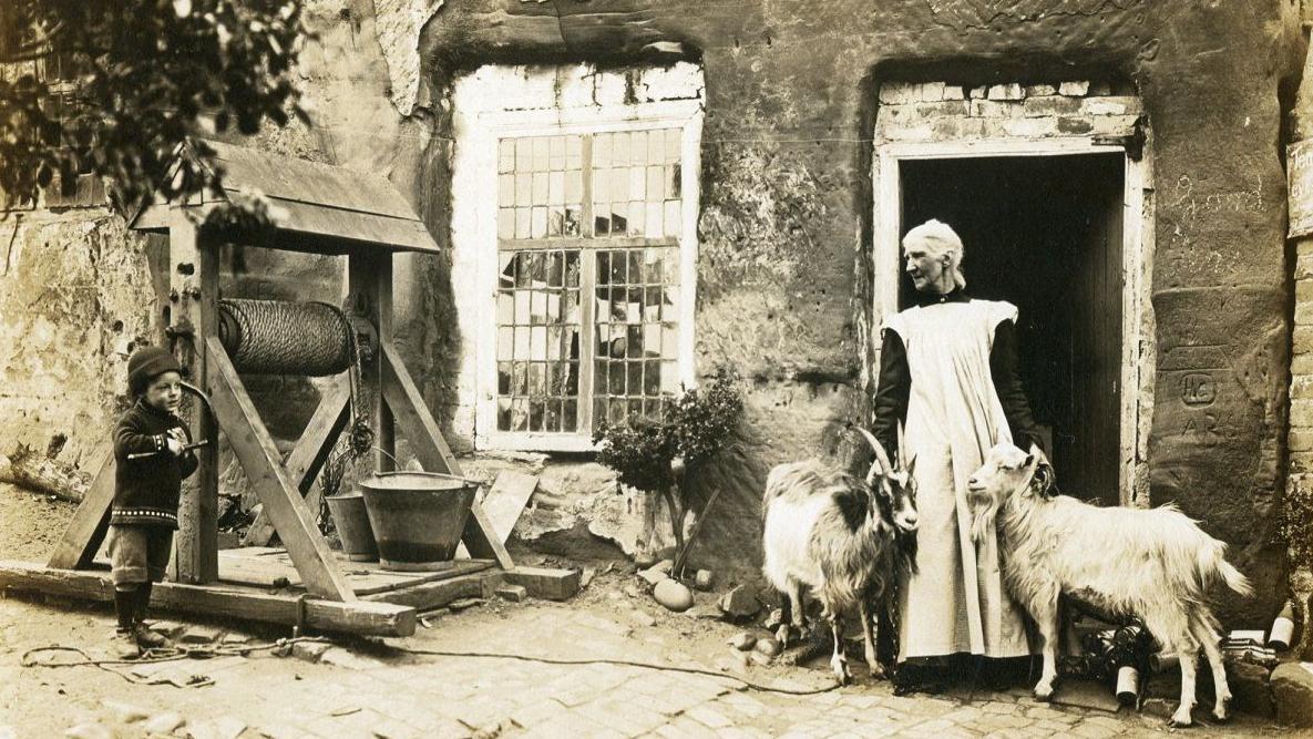 Uma mulher vestindo um casaco branco com mangas pretas está ao lado de uma porta com as mãos sobre uma cabra. Ao lado deles há um poço de madeira coberto com uma corda enrolada e um balde. Ao lado do poço está um menino de chapéu, moletom e calça. 