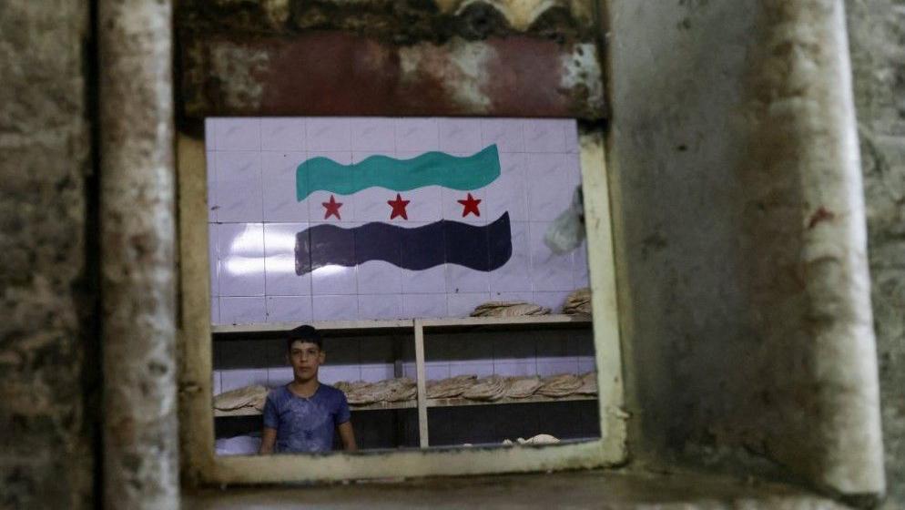 عامل يقف في مخبز وخلفه علم (الثورة) السورية، بعد الإطاحة بالرئيس السوري بشار الأسد، في العاصمة السورية دمشق يوم 6 يناير/كانون الثاني 2025.