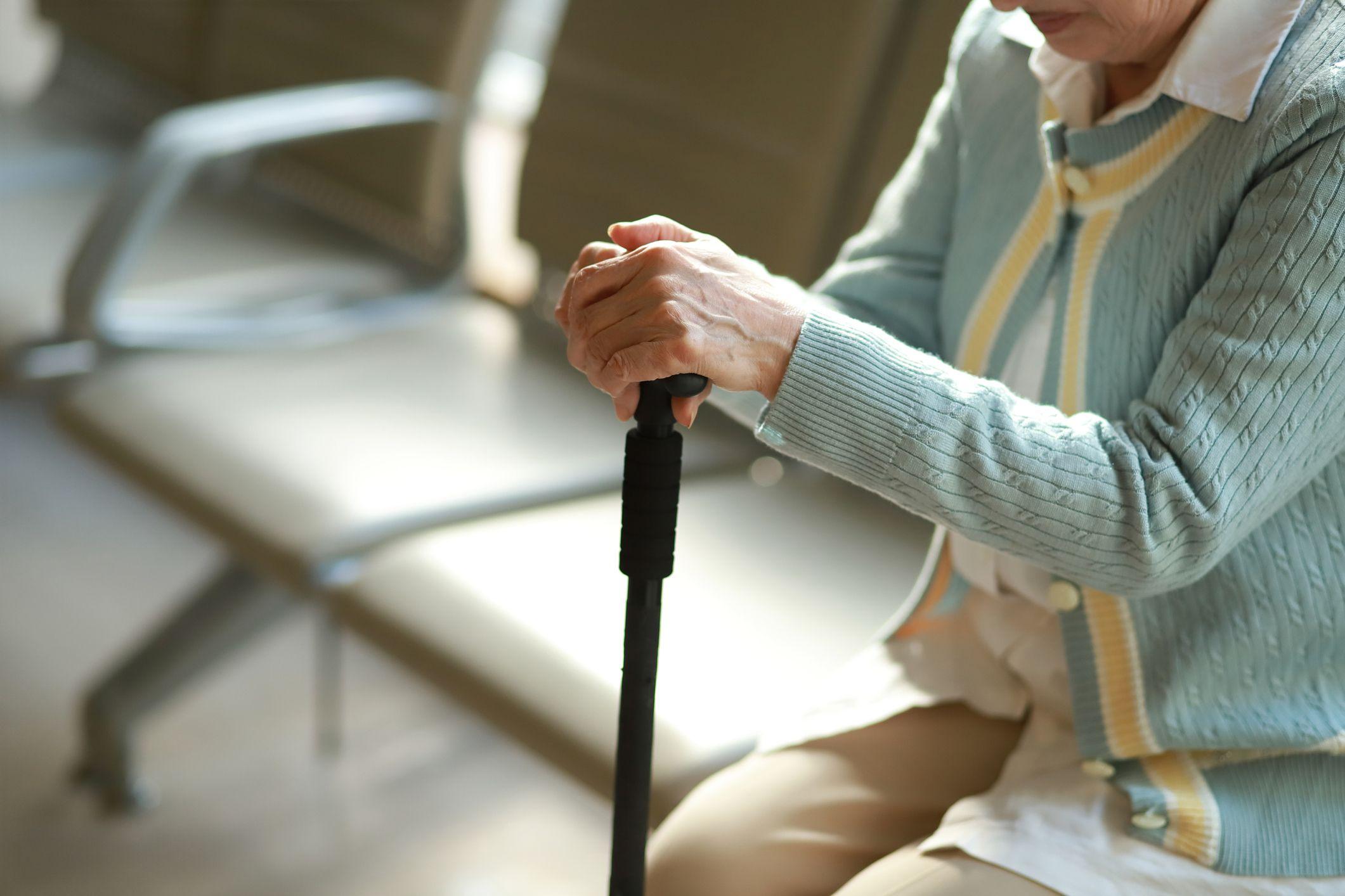 Mulher idosa sentada em sala de espera de hospital segurando uma bengala.