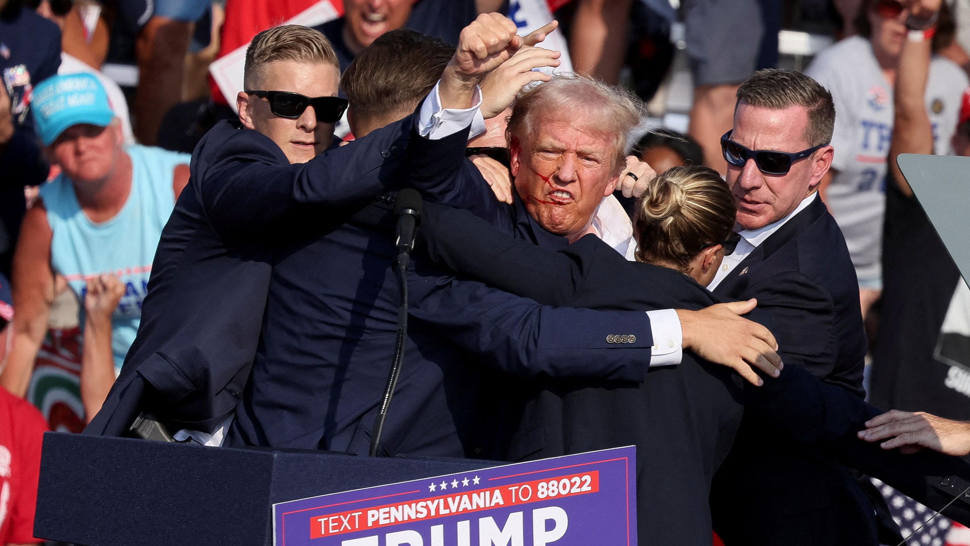 Trump, ainda no palco, rodeado de agentes da segurança, com a mão em punho para o alto e o lado direito do rosto com sangue. 