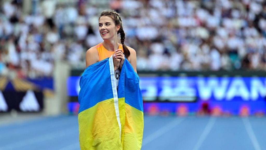 Yaroslava Mahuchikh na pista de atletismo abraçando a bandeira ucraniana