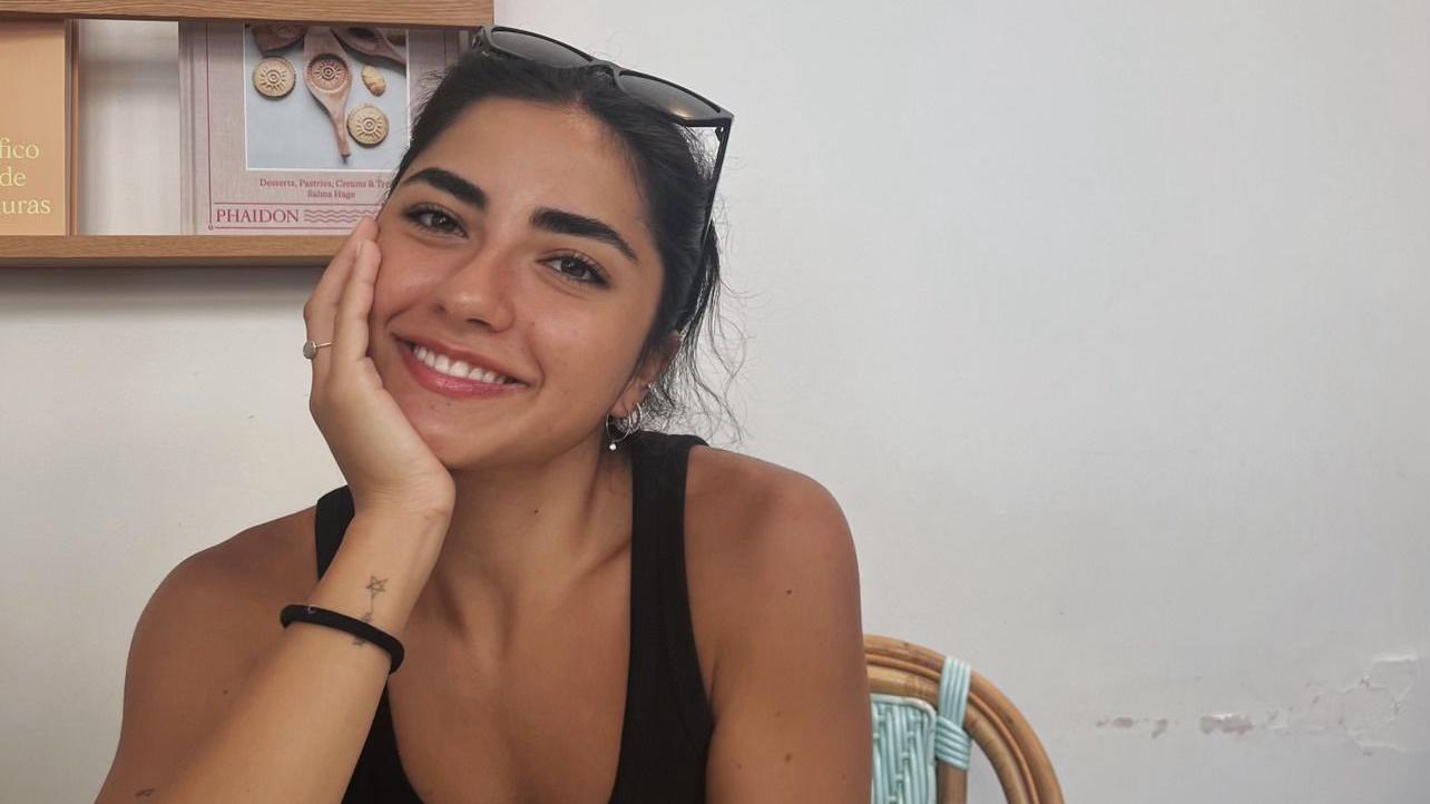 A foto mostra Mary, uma garota com cabelos escuros, presos sorrindo para a câmera. Ela está usando óculos escuros na cabeça e uma blusa preta, sentada em uma cadeira. Ao fundo, há uma estante com livros.