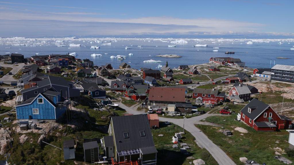 Um aglomerado de casas, algumas pintadas de vermelho e azul, cercadas por grama e conectadas por estradas pavimentadas, em frente ao mar pontilhadas de icebergs na Baía de Disko, 15 de julho de 2024 em Ilulissat, Groenlândia