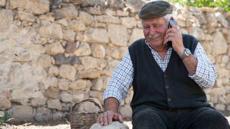 Homem idoso sentado em mureta ao ar livre falando ao celular na Sardenha