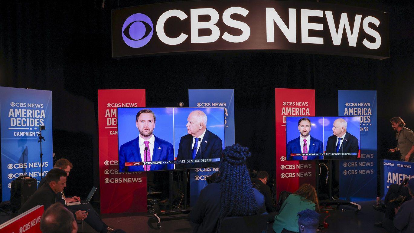 A foto mostra pessoas em um estúdio, de costas, assistindo ao debate por meio de duas televisões. 