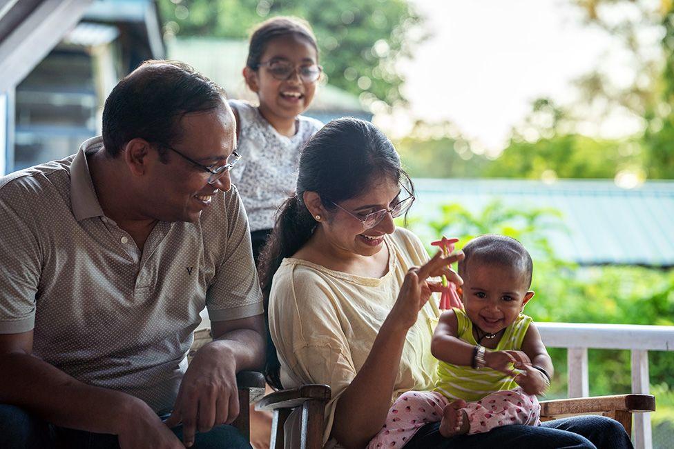 Gaurav dan istrinya, keduanya mengenakan kacamata dan tersenyum, menggendong bayi perempuan mereka yang mengenakan rompi hijau limau dan celana pink. Putri mereka yang lebih tua, mengenakan kaos hitam putih, berdiri di belakang, juga tersenyum.