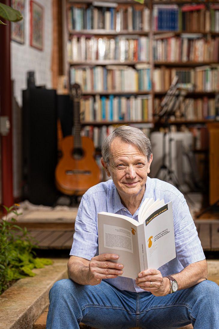 Foto de André Singer sentado com um livro nas mãos, olhando para a câmera.