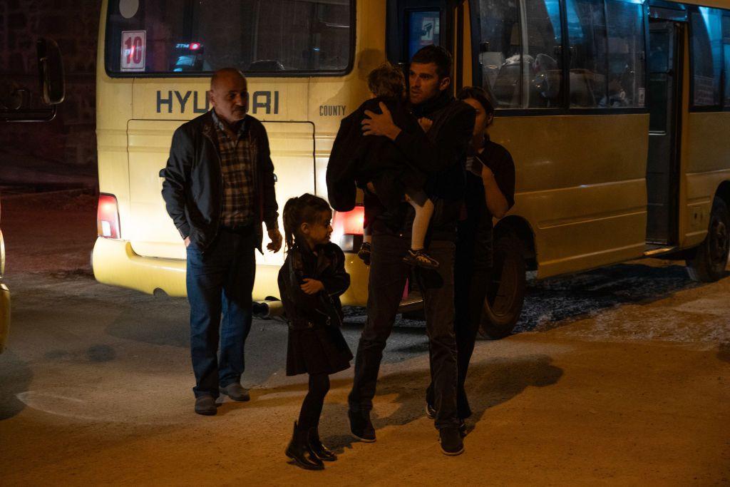 Un hombre con un niño en sus brazos, una niña y otros dos adultos detrás de un autobús que traslada a los refugiados.