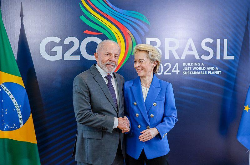 Lula usando terno cinza escuro ao lado de Ursula Von der Leyen usando terno azul fazendo um aperto de mãos