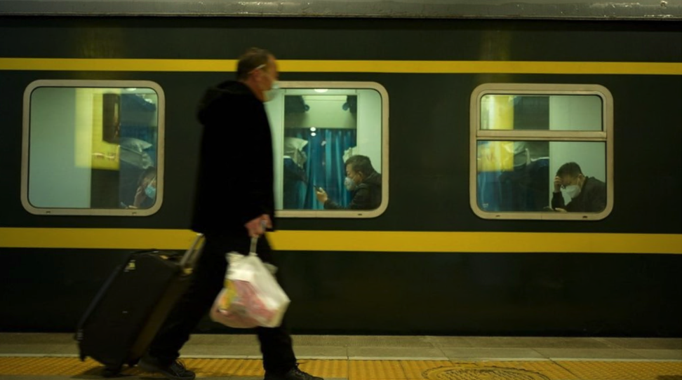 A man carrying a bag and dragging a suitcase, wearing a mask, walks past a train with three men at three windows also wearing masks inside