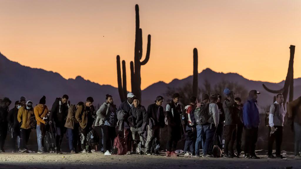 Uma fila de migrantes, alguns com casacos e chapéus quentes, é vista em frente a cactos e montanhas em um centro de processamento da Patrulha de Fronteira dos EUA após cruzar a fronteira EUA-México em 7 de dezembro de 2023 em Lukeville, Arizona