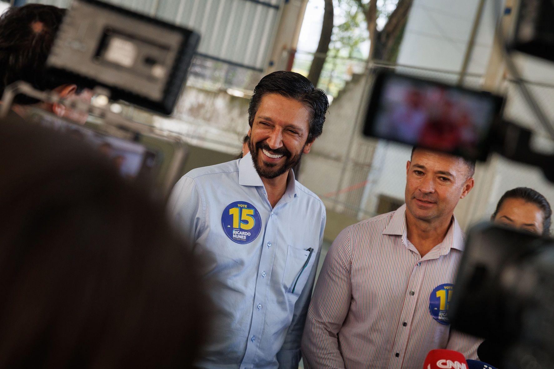 Nunes e Mello Araújo sorrindo, rodeados por jornalistas e câmeras