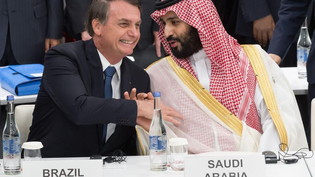 Sentados diante de placas com os nomes de seus respectivos países, Jair Bolsonaro e Mohammed bin Salman sorriem enquanto apertam as mãos