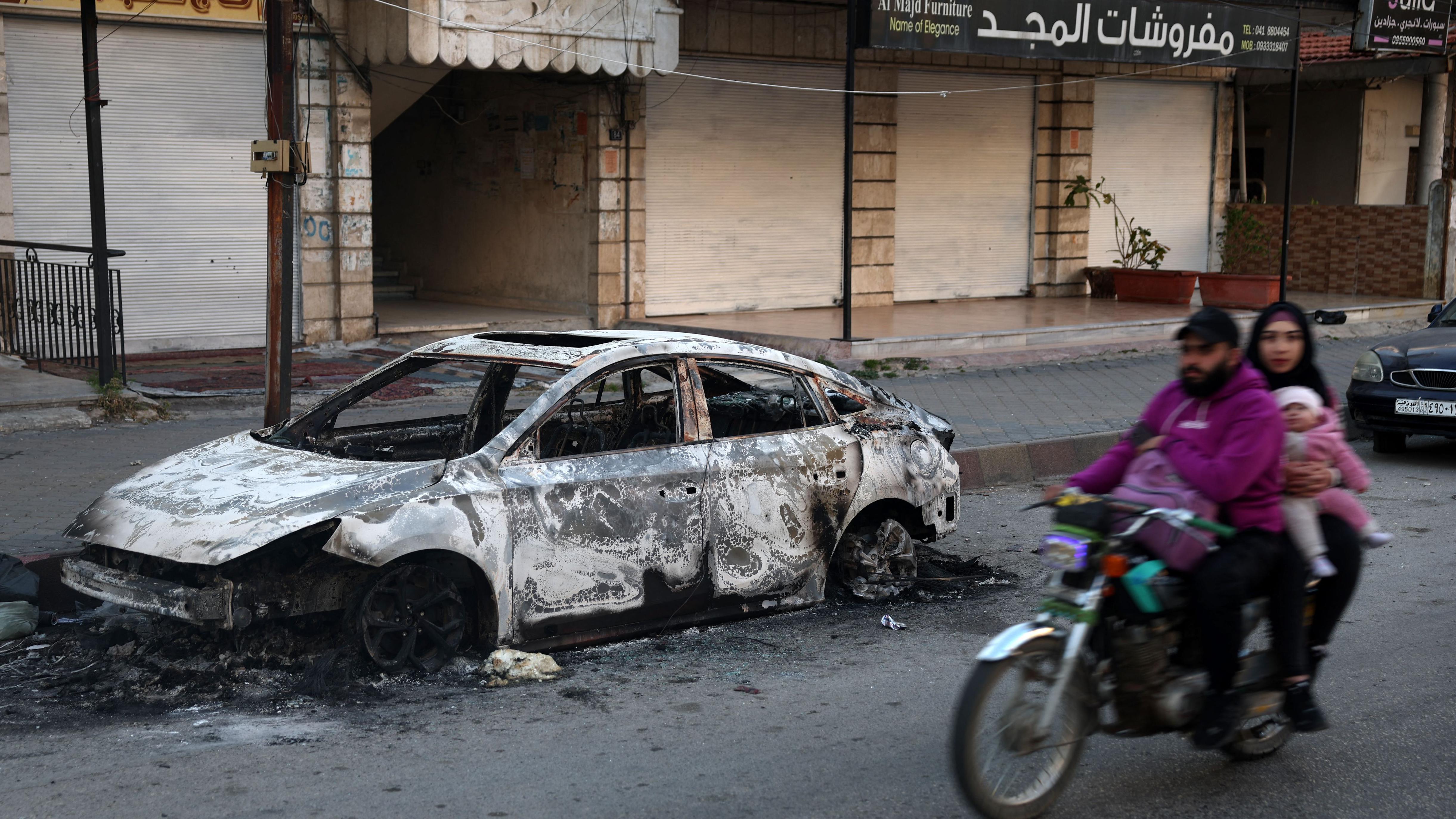 أفراد عائلة يركبون دراجة نارية يمرون بسيارة متفحمة في بلدة جبلة في محافظة اللاذقية الساحلية السورية في 12 مارس/آذار 2025