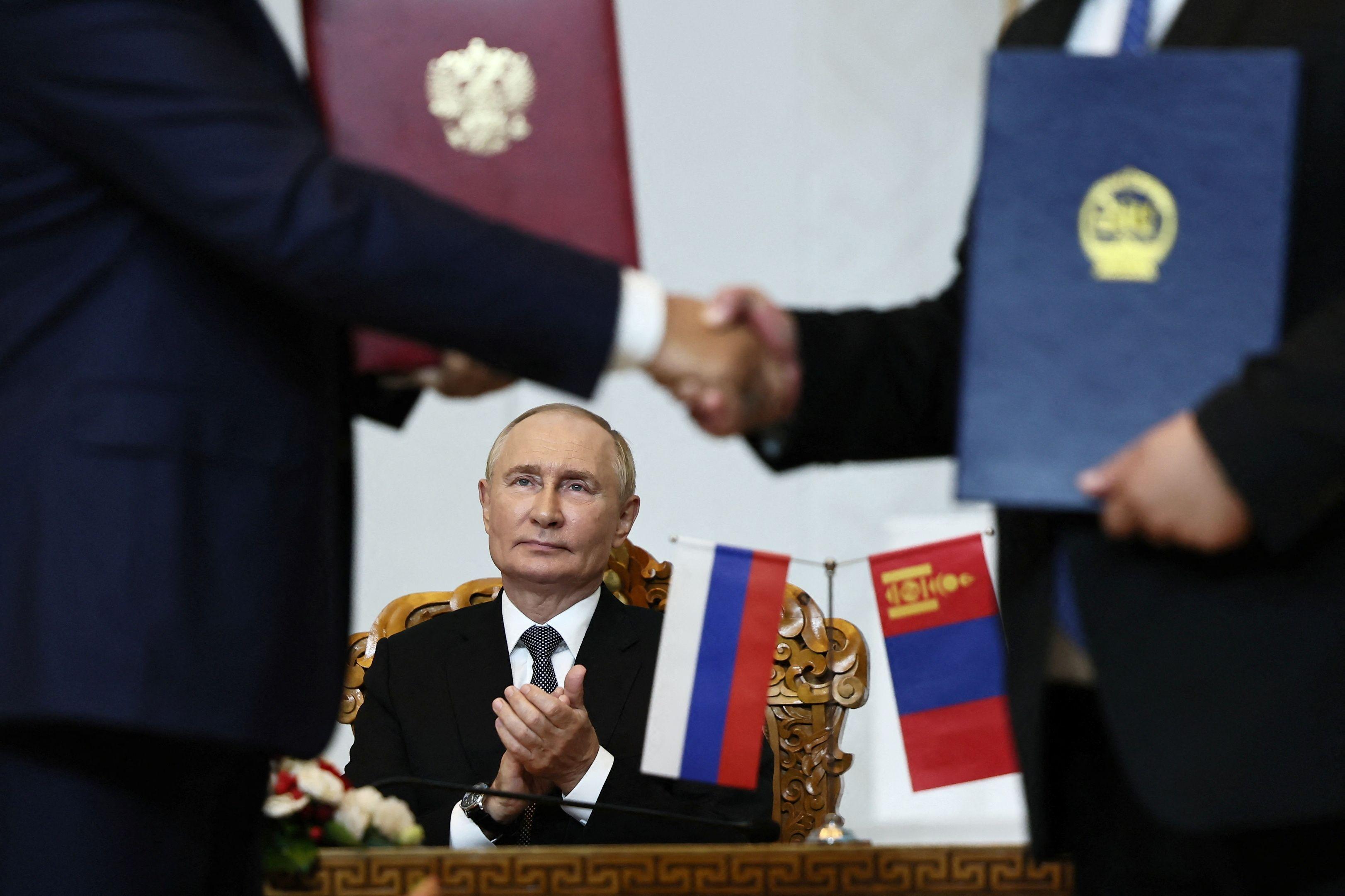 Putin sentado em frente a uma mesa, enquanto duas pessoas apertam as mãos na frente dele.