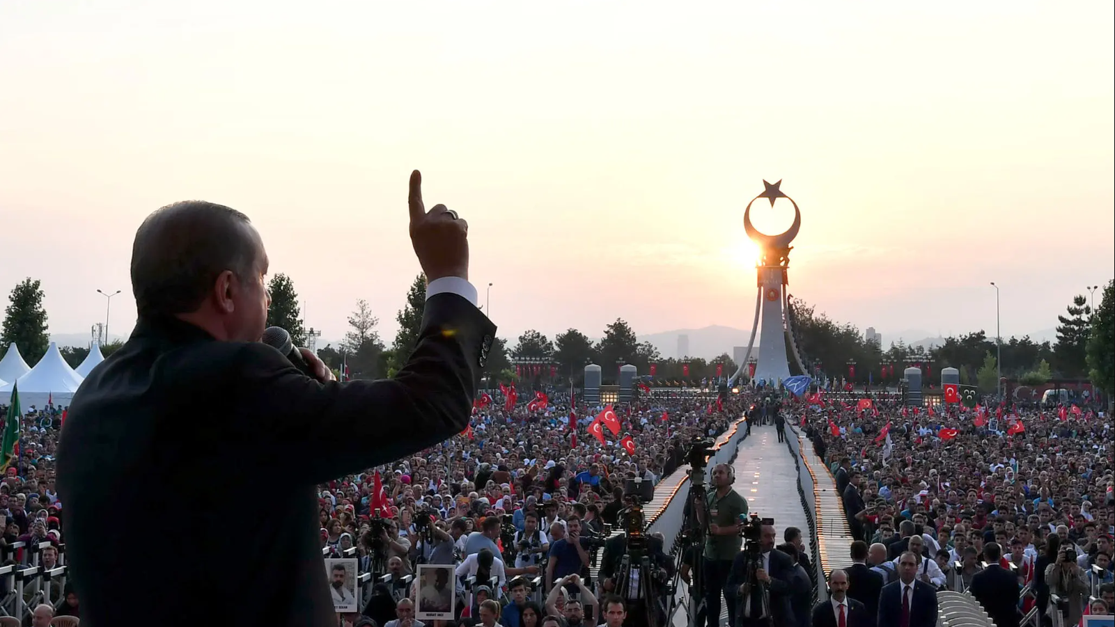 أردوغان بلباس رسمي يرفع سبابته عالياً وأمامه حشد جماهيري يحمل عدد منهم العلم التركي