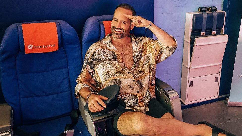 Jay Robert, de camisa estampada, short e sandália, sentado em um assento de avião, fazendo saudação enquanto sorri