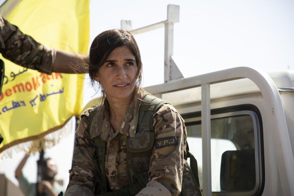 الصورة لشابة مقاتلة في صوف قسد ببزة عسكرية وفي الخلفية العلم الكردي الأصفر