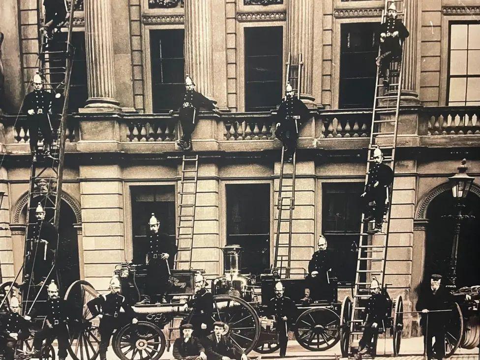 Bomberos posando con sus carros y escaleras frente a un edificio 